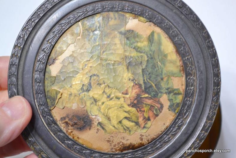 Op de afbeelding: Een ronde, antieke metalen lijst met een bloemmotief. De ingelijste afbeelding toont figuren in gele en rode kleding, tegen een achtergrond van groen gebladerte. De afbeelding heeft een verweerde, verouderde uitstraling.