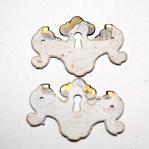 May include: Two antique keyhole escutcheons with a white painted finish and gold accents. The escutcheons are in the shape of a stylized floral design.