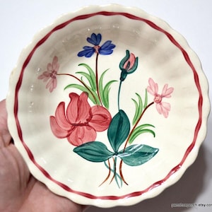 May include: A small, off-white ceramic dish with a scalloped edge and a red border. The dish is decorated with hand-painted flowers in shades of red, pink, blue, and green. The flowers are arranged in a floral pattern.