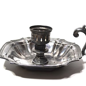 May include: A silver-toned candle holder with a decorative handle. The candle holder has a scalloped dish base and a central cup with decorative cutouts. The handle curves upwards. The item is on a white background.