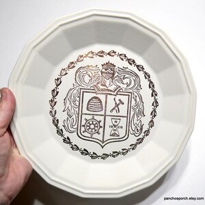 May include: Cream-coloured, octagonal plate with a brown crest design. The crest features a beehive, crossed tools, a ship's wheel, and an hourglass, surrounded by a laurel wreath. The plate is held by a hand.