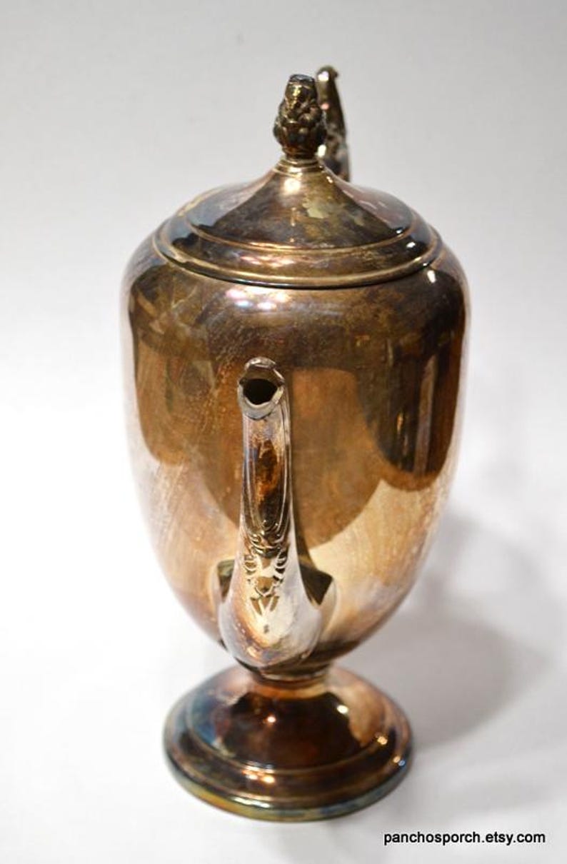 Pu&ograve; includere: Una caffettiera in argento placcato con un manico decorativo e un coperchio con un pomo. La caffettiera ha un corpo rotondo e una base svasata.