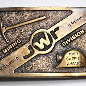 May include: A rectangular, bronze-coloured belt buckle with the text "JWf" in the centre. The buckle features a pickaxe, a hard hat, and the words "Mining," "Brookwood," "Alabama," "Division," and "1983 Safety Award."
