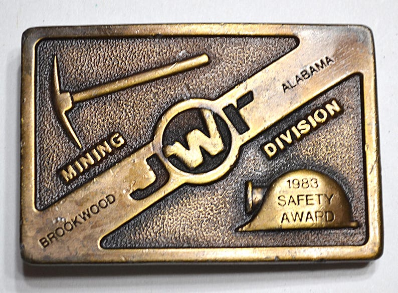 May include: A rectangular, bronze-coloured belt buckle with the text "JWf" in the centre. The buckle features a pickaxe, a hard hat, and the words "Mining," "Brookwood," "Alabama," "Division," and "1983 Safety Award."