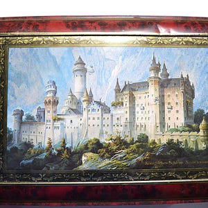 May include: A framed print of Neuschwanstein Castle, featuring a detailed illustration of the castle with its towers and turrets set against a mountain backdrop. The frame is a dark red with a decorative gold border.