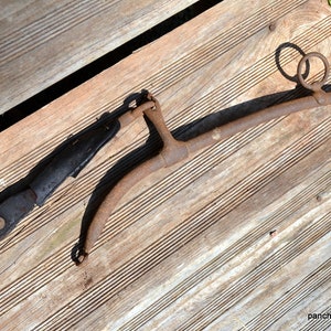 May include: An antique horse harness yoke and leather strap, resting on weathered wooden planks. The yoke is made of rusted metal with two rings and a curved design. The leather strap is black with metal hardware.