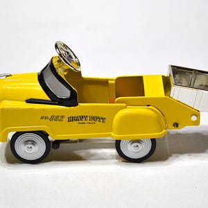 May include: A yellow toy dump truck with black and silver details. The truck features a steering wheel, a dump bed, and the text "No. 882 Heavy Duty Dump Truck". The wheels are black with silver rims.