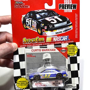 May include: A 1995 Racing Champions NASCAR die-cast stock car, featuring a blue and yellow design with the number 51. The packaging includes a collector's card and display stand, with the name Curtis Markham. The card reads "1995 Preview Edition."