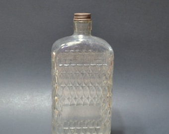 May include: A clear glass bottle with a rectangular body featuring a textured diamond pattern on the sides. The bottle has a narrow neck and a small, metallic, ribbed screw-top cap. The glass is transparent, showing the light reflecting off the facets.