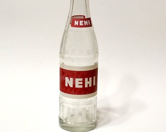 May include: Clear glass bottle with a textured design and red and white labels. The labels read "NEHI". The bottle is empty and stands upright against a white background. The bottle is a vintage soda bottle.