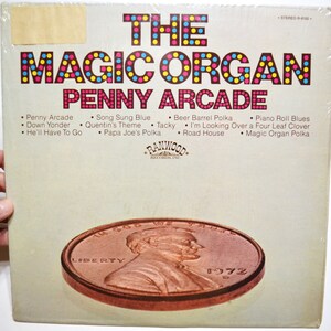 May include: A vintage vinyl record album cover titled The Magic Organ Penny Arcade by Ranwood Records. The cover features a large copper-colored penny graphic at the bottom and a tracklist including Penny Arcade, Song Sung Blue, and Beer Barrel Polka printed in brown text against a light beige background.