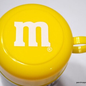 May include: A yellow mug with a white "M" logo on the top. The mug has a handle and a glossy finish. The mug is a promotional item for M&M's candy.