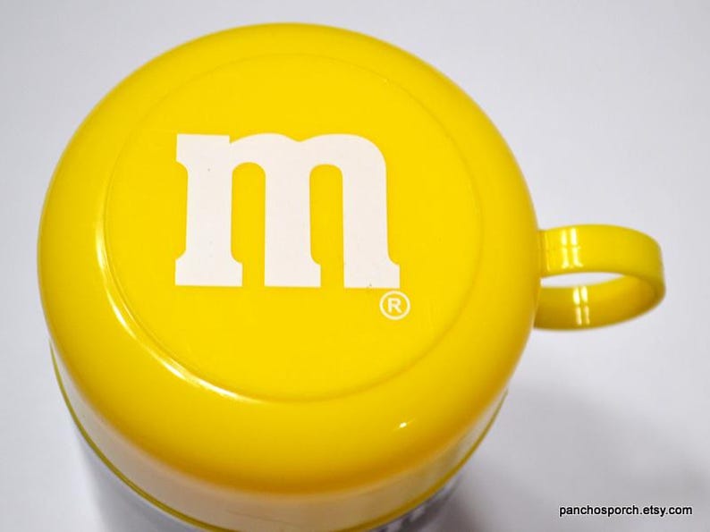 May include: A yellow mug with a white "M" logo on the top. The mug has a handle and a glossy finish. The mug is a promotional item for M&M's candy.