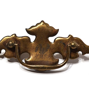 May include: Antique brass drawer pull with a decorative design. The pull features a central ornate shape with curved wings on either side and a curved handle. The hardware is mounted with two screws.