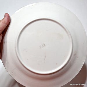 Op de afbeelding: Een witte keramische plaat met een licht getextureerd oppervlak. De plaat heeft een brede rand en een centraal verzonken gebied. Er zijn kleine onvolkomenheden en markeringen op het oppervlak. De plaat wordt door een hand vastgehouden.