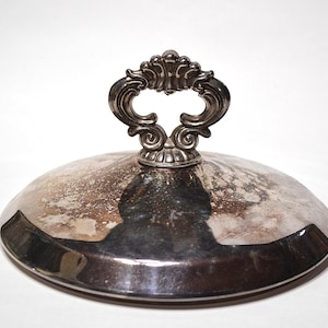 May include: A silver-coloured, domed lid with an ornate handle. The handle has a decorative, open design. The lid has a reflective surface with some wear and discolouration, suggesting age. The item is likely a serving dish lid.