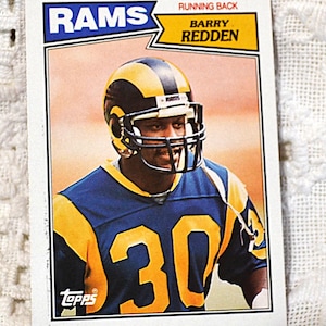May include: A vintage Topps trading card featuring a football player wearing a blue and yellow Rams jersey with the number 30. The card has the text "RAMS" in blue and "RUNNING BACK BARRY REDDEN" in black.