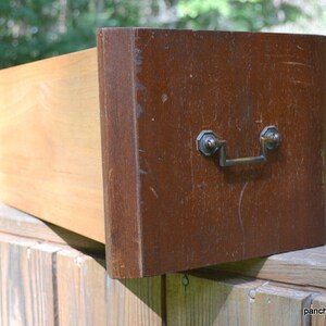 May include: An open wooden drawer with a dark brown front and a light brown interior. The drawer has a metal handle and is set against a natural outdoor background. The drawer is likely from a dresser or cabinet.