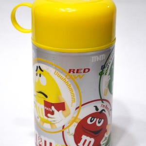 May include: A yellow and silver M&M's thermos with a yellow lid and handle. The thermos features cartoon M&M's characters and the text "RED" and "MATH". The thermos is cylindrical and has a silver body.