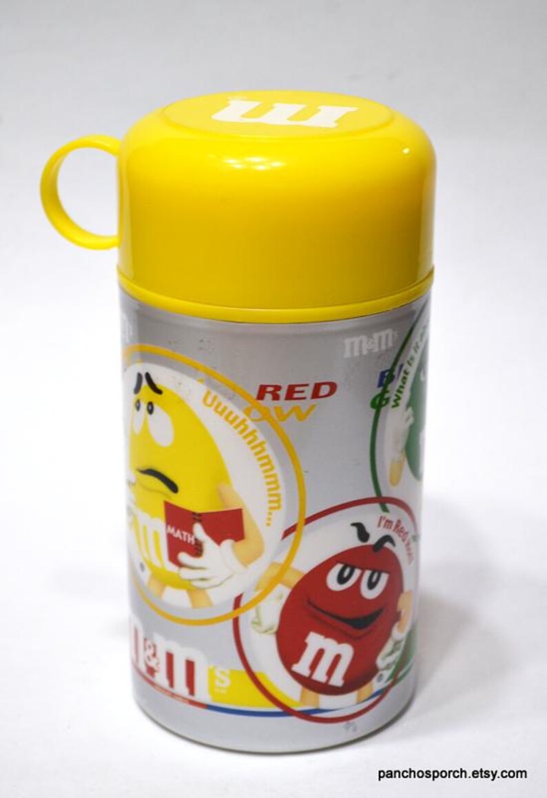 May include: A yellow and silver M&M's thermos with a yellow lid and handle. The thermos features cartoon M&M's characters and the text "RED" and "MATH". The thermos is cylindrical and has a silver body.