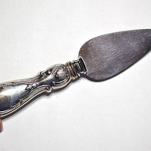 May include: Silver cheese knife with a decorative handle and a wide, flat blade. The handle features ornate detailing, and the blade has a slightly textured surface. A classic serving utensil.