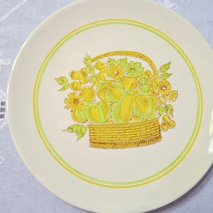May include: Off-white ceramic plate with a yellow and green fruit basket design. The basket is filled with yellow and green fruits and flowers. The plate has a yellow and green border.