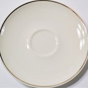 May include: Cream-colored saucer with a gold rim. The saucer has a slightly concave center designed to hold a teacup. The gold rim adds a touch of elegance to the simple, classic design. The saucer is made of ceramic.
