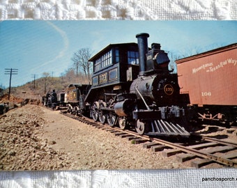 May include: A vintage postcard featuring a black steam locomotive numbered 173 on railroad tracks. A brown freight car labeled Missouri-Kansas-Texas Rail Way 106 is attached. The scene includes a rocky landscape, utility poles, and a clear blue sky.