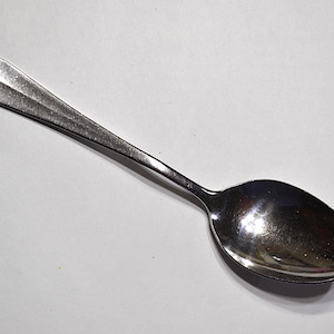 May include: A stainless steel spoon with a rounded bowl and a long, slender handle. The handle has a subtle, ridged design. The spoon is set against a plain white background, highlighting its polished, reflective surface. This is a common utensil for eating.
