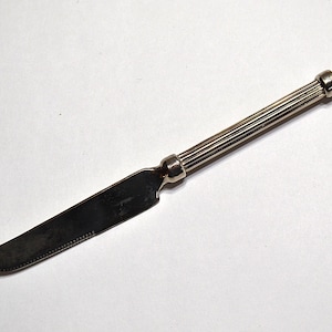 May include: A silver cheese knife with a curved blade and a ridged handle. The knife has a pointed tip and a small, curved end. The handle is cylindrical with a rounded end. The knife is on a white background.