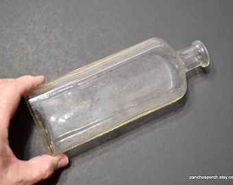 May include: A clear, rectangular glass bottle with a rounded neck and a slightly yellowed rim. The bottle is held by a hand and appears to be an antique or vintage item. The bottle's surface shows signs of age and wear.
