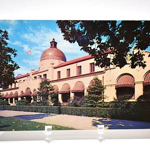 May include: Vintage postcard featuring a beige building with a red-tiled dome and awnings. The building has arched windows and is surrounded by trees and a manicured lawn. The sky is blue with clouds.