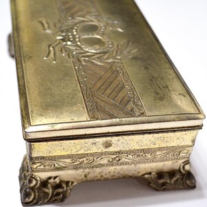 May include: An antique brass box with intricate engravings. The lid displays an engraved design with a central oval and ribbon motif, surrounded by floral patterns. The box is supported by decorative, claw-like feet, showing an aged patina.
