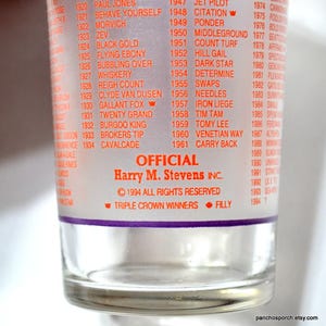 Puede incluir: Vaso transparente con una lista de ganadores de carreras de caballos en texto rojo. El vaso presenta el texto "OFFICIAL Harry M. Stevens INC." y "&copy; 1994 TODOS LOS DERECHOS RESERVADOS". La parte inferior del vaso dice "TRIPLE CROWN WINNERS &bull; FILLY".