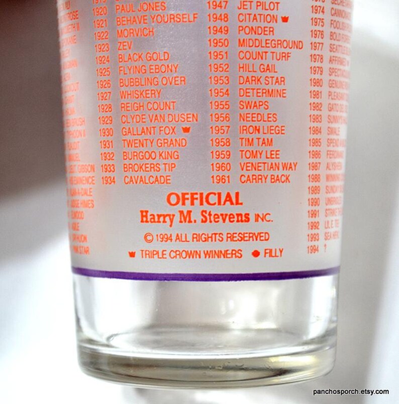 Puede incluir: Vaso transparente con una lista de ganadores de carreras de caballos en texto rojo. El vaso presenta el texto "OFFICIAL Harry M. Stevens INC." y "&copy; 1994 TODOS LOS DERECHOS RESERVADOS". La parte inferior del vaso dice "TRIPLE CROWN WINNERS &bull; FILLY".