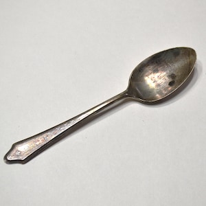 May include: A silver-toned metal spoon featuring a tarnished finish and an elongated oval bowl. The handle is tapered with a pointed end and displays a subtle, embossed floral pattern along its length, showing signs of age and oxidation.