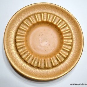 May include: A round, golden-toned ceramic dish featuring a central basin surrounded by a wide, fluted rim. The surface displays a textured, metallic gold finish over a lighter base, with a repeating scalloped pattern embossed along the outer edge.