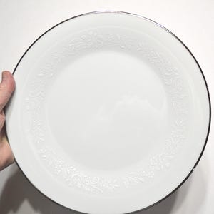 May include: A round white dinner plate featuring a delicate embossed floral pattern around the rim and a thin silver metallic band along the outer edge. The center of the plate is smooth and plain, providing a clean surface for food presentation.