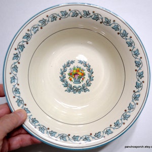 May include: Cream-colored ceramic bowl with a teal blue rim and a decorative floral border. The center features a basket of colorful fruit surrounded by a laurel wreath. A classic design for serving.
