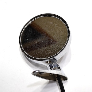May include: A chrome side-view mirror with a round, slightly convex mirror surface. The mirror has a shiny, reflective finish and a black wire extending from the base. The mirror is likely for a classic car.
