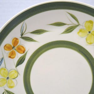 May include: A white ceramic saucer featuring a wide olive green band along the outer rim and a thinner green circle in the center. The surface is decorated with hand-painted yellow and orange four-petaled flowers and green leaves.