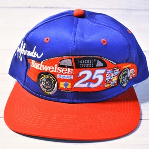 May include: A blue baseball cap with a red brim and a top button. The front features an embroidered orange race car with the number 25, the Budweiser logo, and GMAC text. The name Ken Schrader is embroidered in white on the side of the crown.