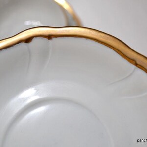 May include: Close-up of white porcelain saucers with a gold rim. The saucers have a scalloped edge and a delicate embossed design. The gold rim adds a touch of elegance to the classic design.