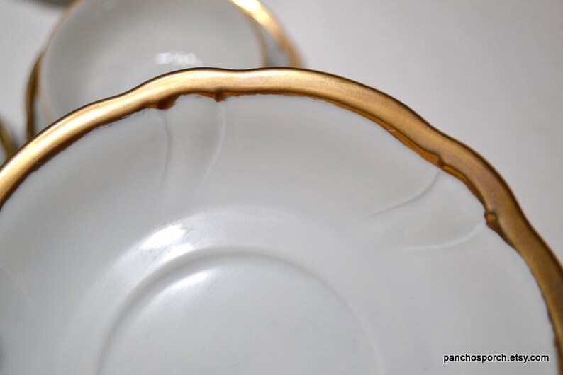 May include: Close-up of white porcelain saucers with a gold rim. The saucers have a scalloped edge and a delicate embossed design. The gold rim adds a touch of elegance to the classic design.