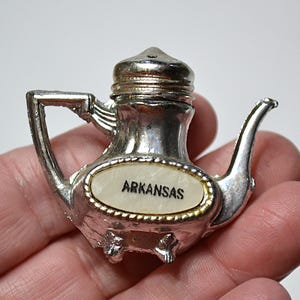 May include: A silver-colored teapot-shaped souvenir with the word "ARKANSAS" on a white oval. The teapot has a handle, spout, and a lid. The souvenir is held in a hand.