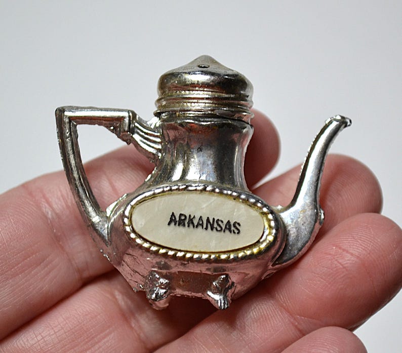 May include: A silver-colored teapot-shaped souvenir with the word "ARKANSAS" on a white oval. The teapot has a handle, spout, and a lid. The souvenir is held in a hand.