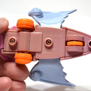 May include: A vintage toy vehicle, featuring a pink body with orange wheels and light blue wings. The underside of the toy is visible, with the text "&copy; 1995 Marvel" and other markings. The toy is held by a person's hand.