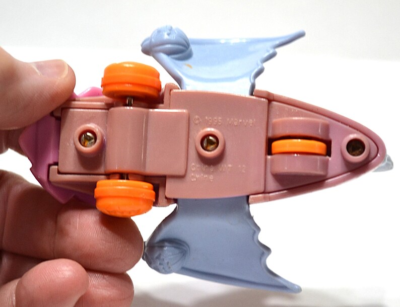 May include: A vintage toy vehicle, featuring a pink body with orange wheels and light blue wings. The underside of the toy is visible, with the text "&copy; 1995 Marvel" and other markings. The toy is held by a person's hand.