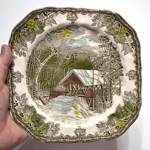May include: A square ceramic plate with scalloped edges featuring a transferware scene of a red covered bridge in a snowy landscape. The border is decorated with embossed green and yellow leaves and brown floral patterns on a cream background.