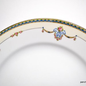 May include: Close-up of a white porcelain plate with a decorative border. The border features a cream-colored band with a blue and gold geometric pattern, pink floral accents, and a blue monogram.